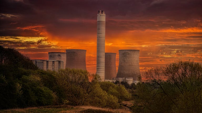 A dramatic sunset casts a warm glow over a power plant, highlighting its industrial structures against the evening sky.