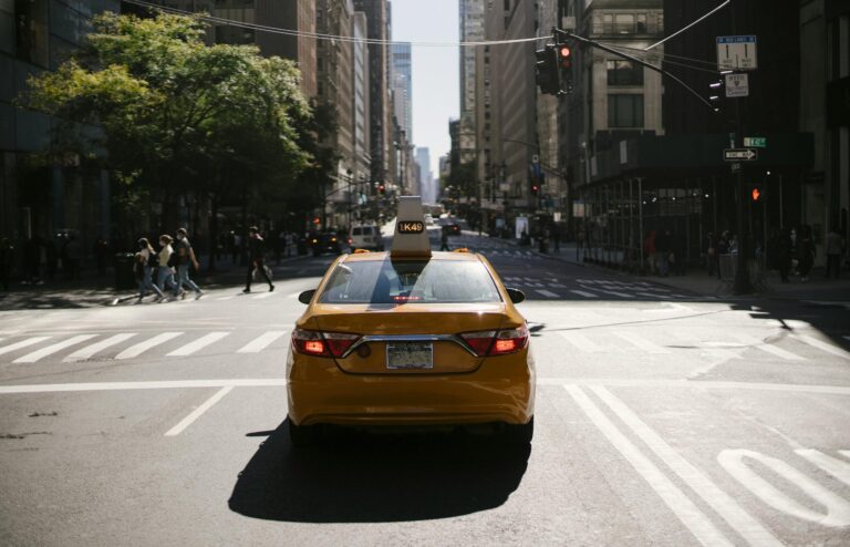 Contemporary taxi with glowing taillights on asphalt road of busy city against crosswalk and traffic light