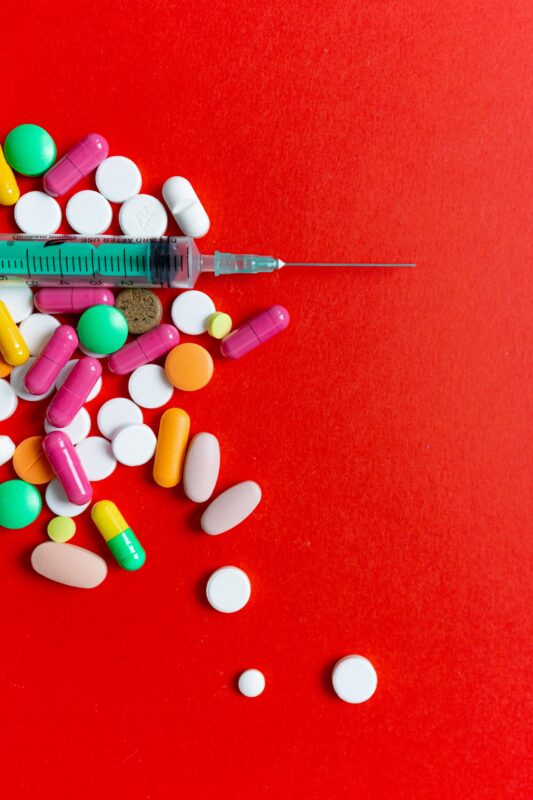 Colorful pills and a syringe on a bright red surface, symbolizing healthcare and medication.