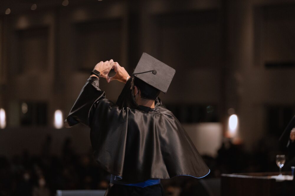 A graduate in cap and gown makes a heart gesture under warm indoor lighting.