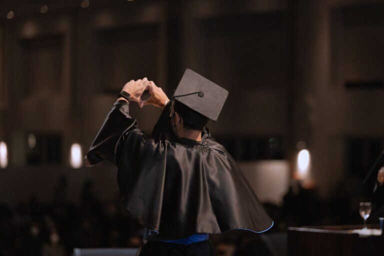 A graduate in cap and gown makes a heart gesture under warm indoor lighting.