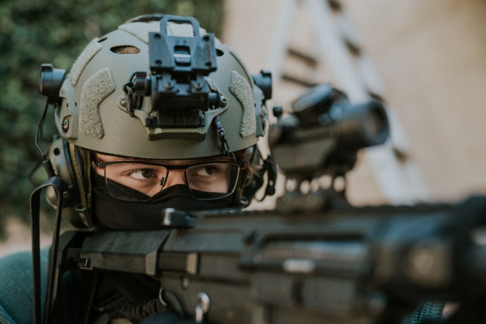 Focused soldier in tactical gear, aiming through optics, military helmet and glasses visible.