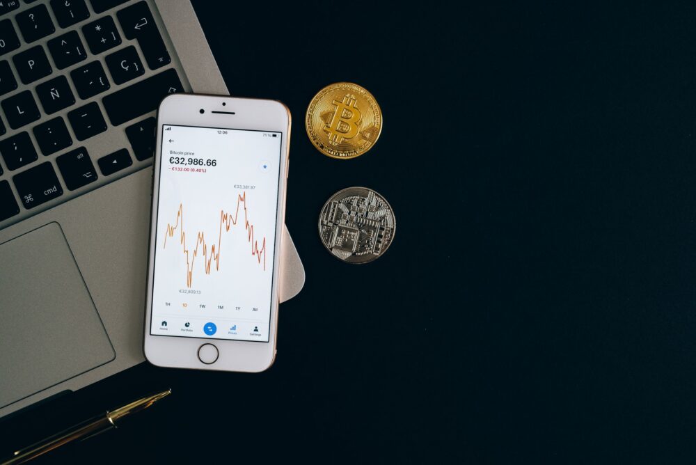 Smartphone displaying Bitcoin price chart alongside Bitcoin and Ethereum coins on black background.